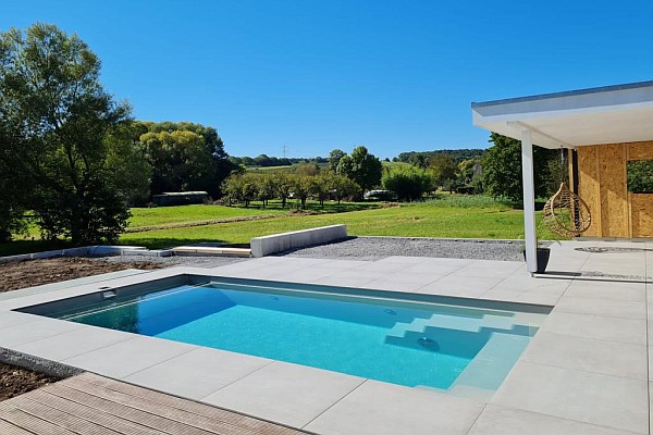 Poolbau bei Heilbronn: Blick auf einen neuen Pool mit blauem Wasser, großen grauen Steinplatten außen, rechts eine Überdachung und im Hintergrund Wiese, Bäume und Hügel