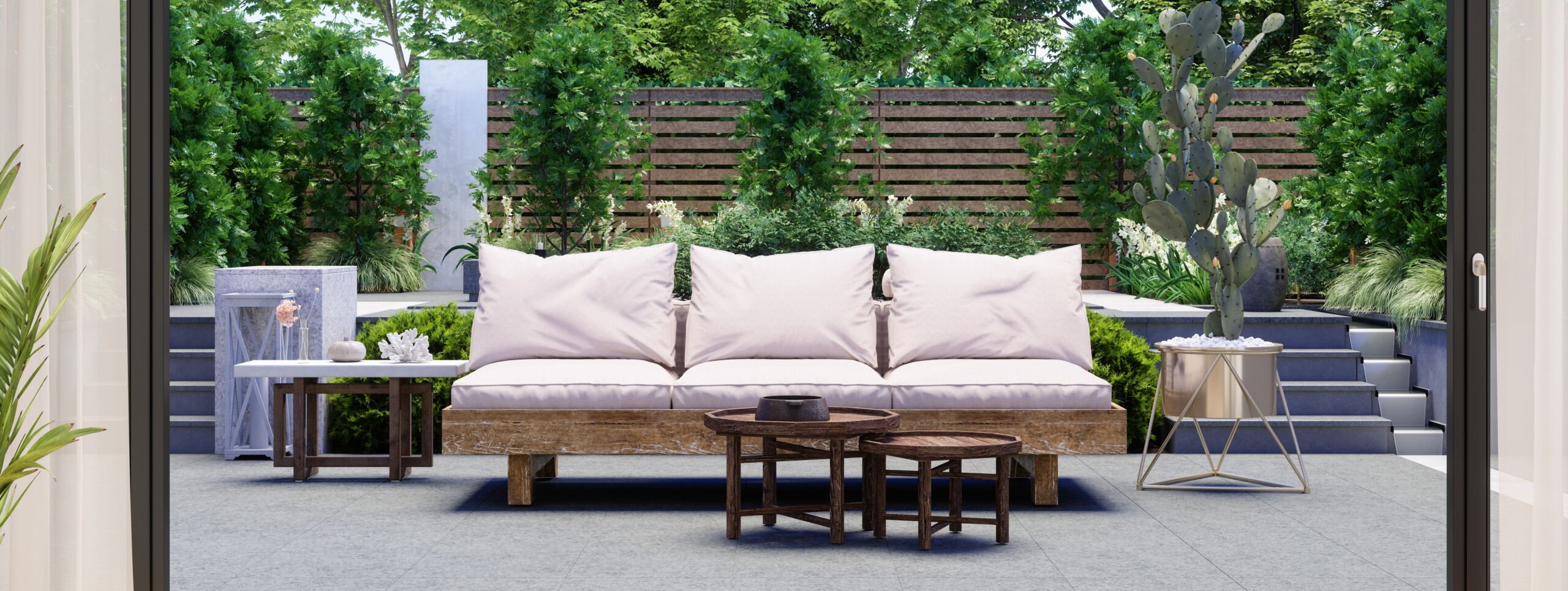 Garten- und Landschaftsbau in Perfektion: Blick aus dem Haus in den modernen Gartenbereich mit bequemen Sofa auf einer Steinterasse mit Treppen zu einer höhergelegenen Ebene mit vielfältiger Bepflanzung bzw. Vegetation