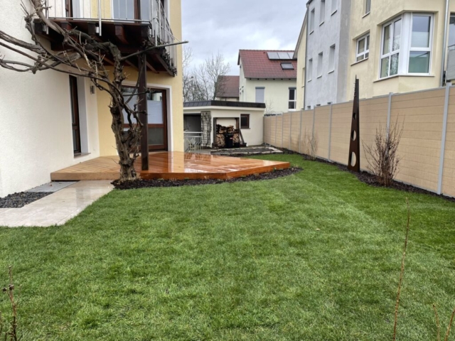 Holzterrasse mit frisch verlegtem Rollrasen, einem beigen Sichtschutz und einem Weg aus Keramikplatten