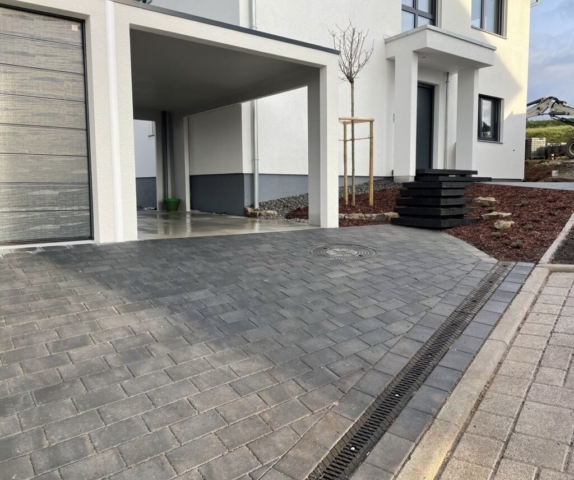 Moderne Einfahrt mit dunkelgrauem Pflaster im Rahmen eines hochwertigen Garten- und Landschaftsbaus. Rechts im Bild führt eine elegante, schwebende Treppe zur Haustür eines weißen, zeitgenössischen Wohnhauses. Ein neu gepflanzter Baum und dekorativer Mulch runden die stilvolle Außenanlage ab.