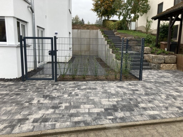 Gartenneuanlage: Bild seitlich von einem Neubeu, mit Treppe zum hinteren Gartenteil, Zaun zur ersten terrasse und gepflasterte Fläche im Vordergrund.