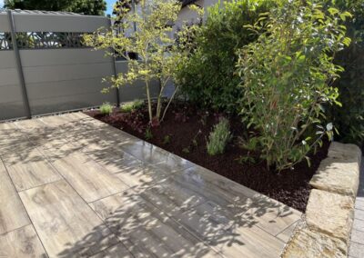 Terrasse mit Holzoptik-Platten, bepflanztem Beet, Sichtschutz, Natursteinmauer und klar gestalteter Außenfläche