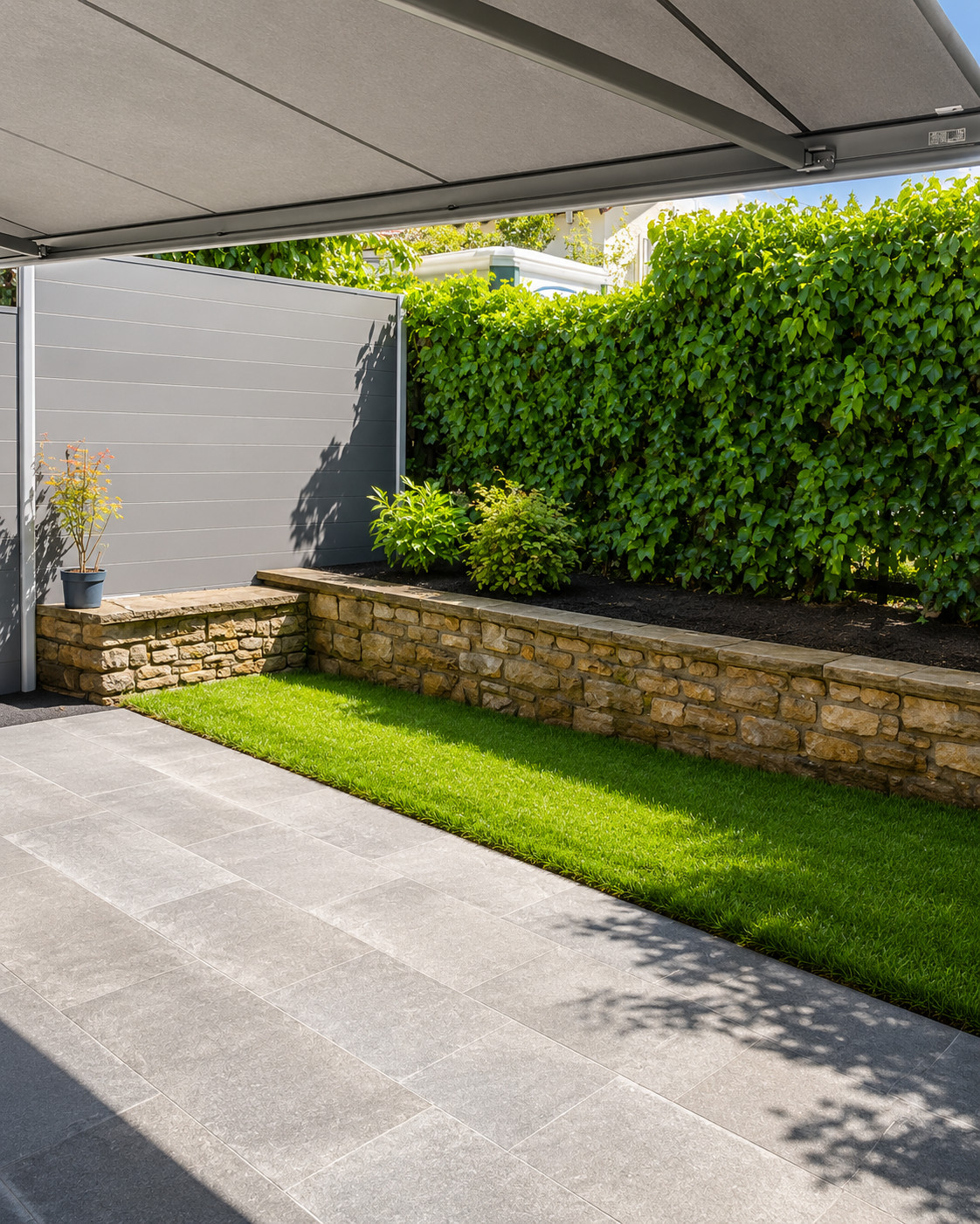 Umbau eines bestehenden Gartens mit Naturstein, Sichtschutz und Bepflanzung Überdachte Terrasse mit grauen Platten, Natursteinmauer, Rasenfläche, Sichtschutz und dichter grüner Bepflanzung