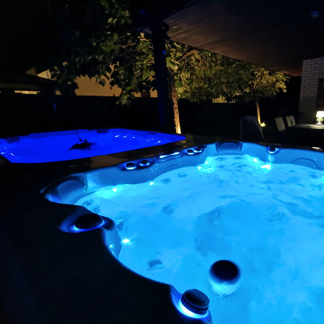 Wellnessgarten Beleuchteter Whirlpool im Garten bei Nacht mit blauem Wasser, überdachtem Bereich und stimmungsvoller Gartenbeleuchtung